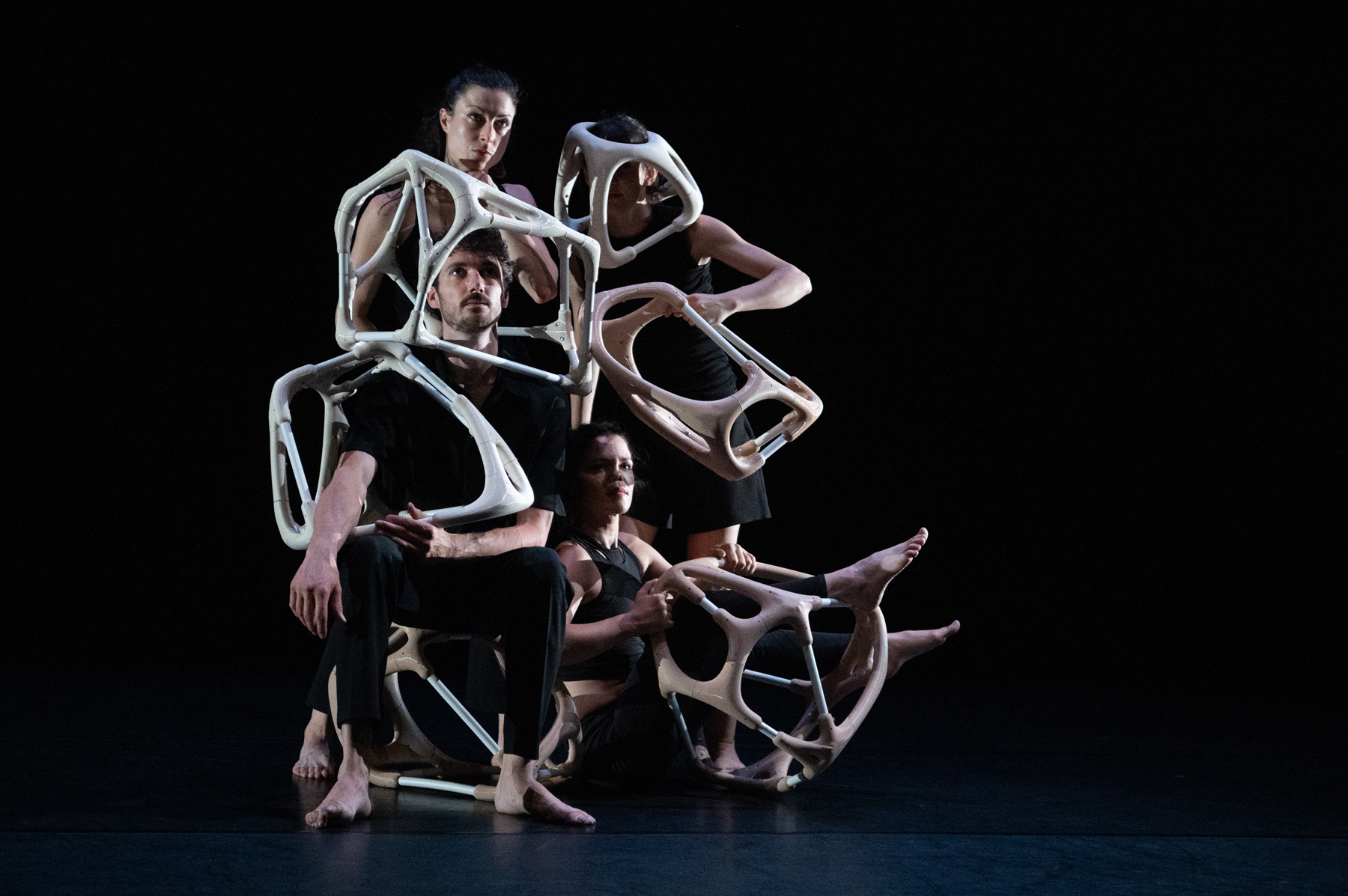 Vêtus de noir, quatre danseureuses accolé·es se tiennent immobiles, le regard rivé au loin. Félix est assis sur une sculpture, Coralie à ses pieds. Derrière, Greta et Sofia sont debout.Iels portent des sculptures volumineuses, aux formes à la fois arrondies et anguleuses, dans des tons beige à blanc cassé. Leur texture évoque celle de l’os ou de la prothèse.L’une des danseureuses a glissé une jambe dans le creux d’une sculpture, l’autre un bras tout entier. Leurs corps imbriqués aux formes osseuses se confondent. Félix et Sofia semblent paré·es de casques d’astronautes.Une lumière spectrale baigne leur pose solennelle.